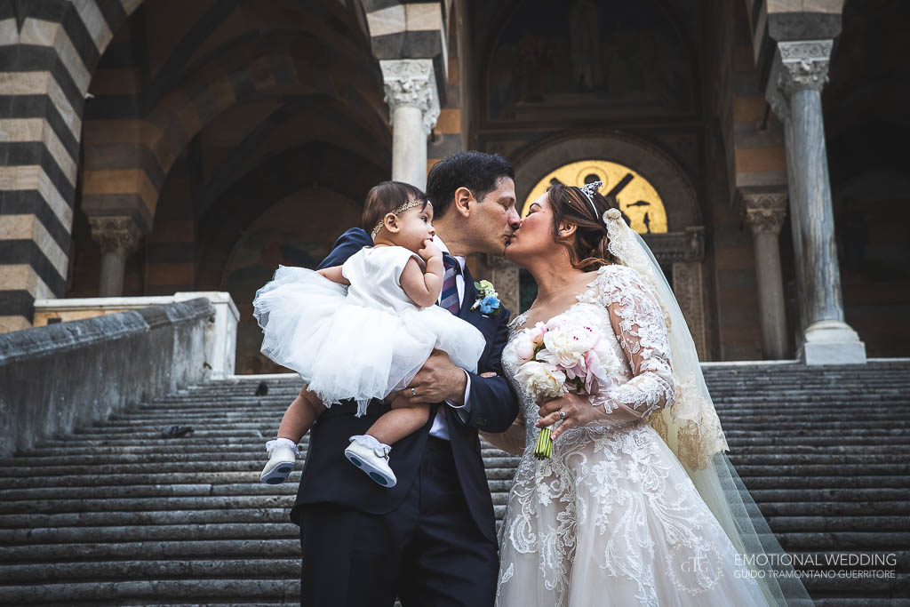 sposi con la figlia in braccio si baciano sulla gradinata del duomo di Amalfi al loro matrimonio