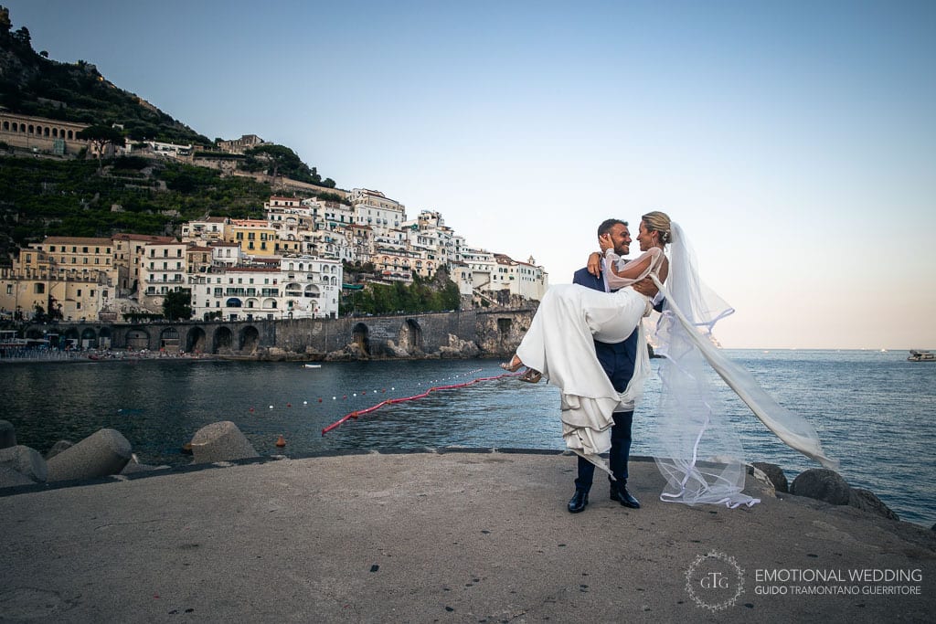 groom takes the bride into his arms on the pier in Amalfi with Atrani in the background