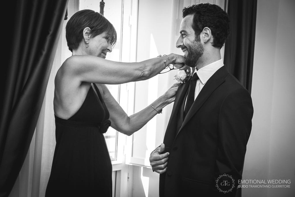 the groom smiles while his mother helps with the boutonniere