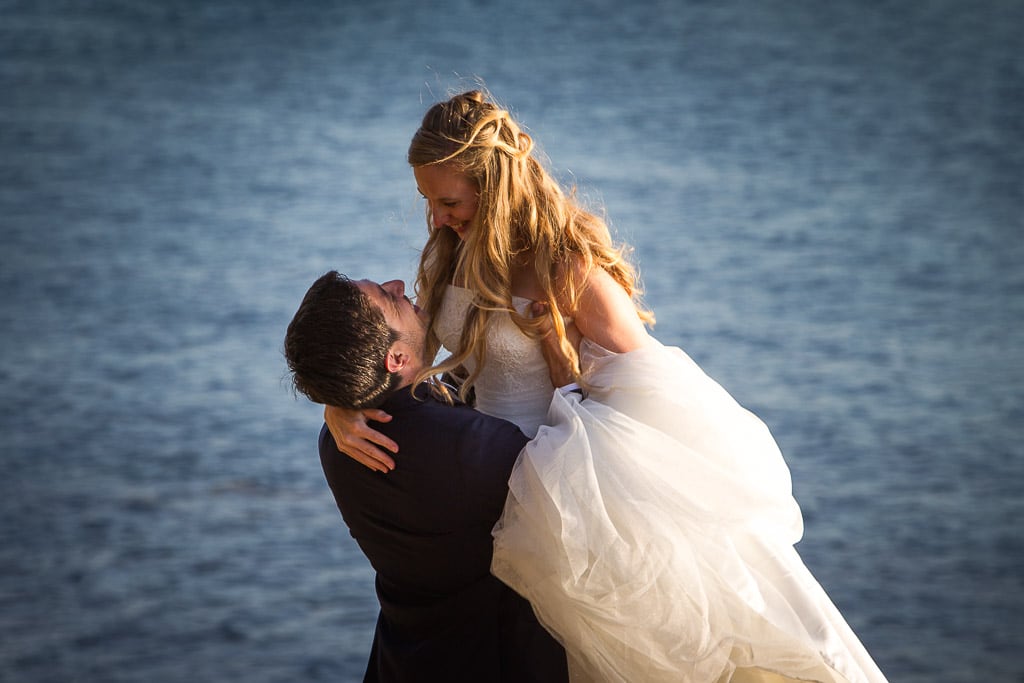 groom holding the bride in his arms and a blue sea in the background