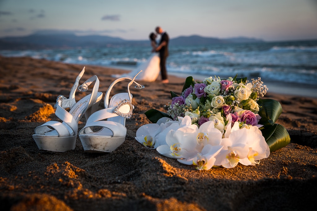 wedding shoes and bouquet on the sand of a beach and a blurred wedding couple in the background