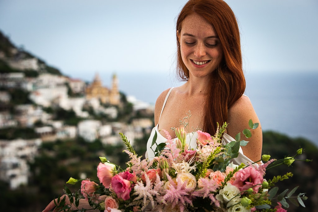bride smiling and holding the bouquet and the town of praiano in amalfi coast in the background