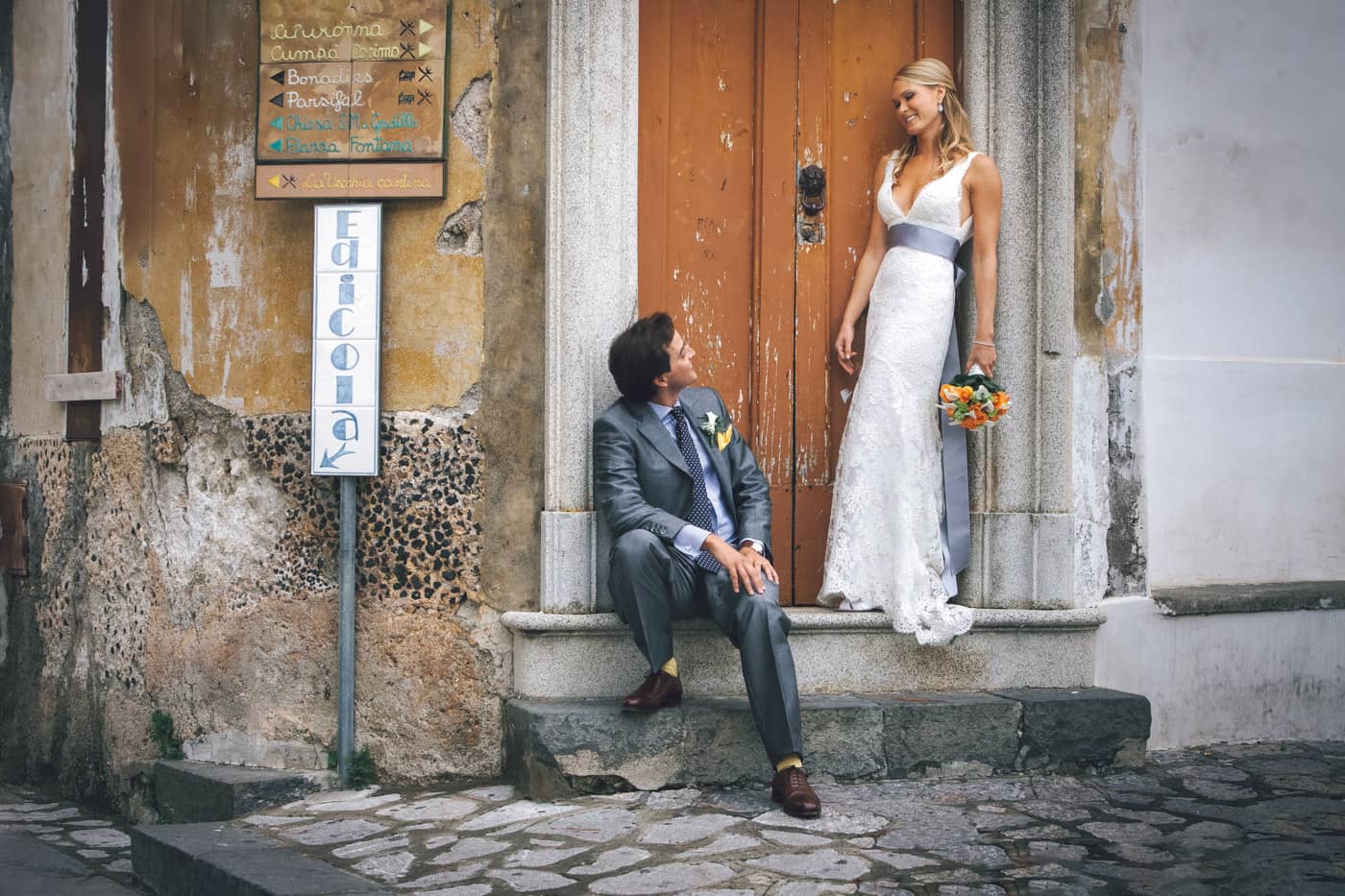 Groom seated looking at his bride in front of an old house door in the main square — Documentary wedding photography Ravello.