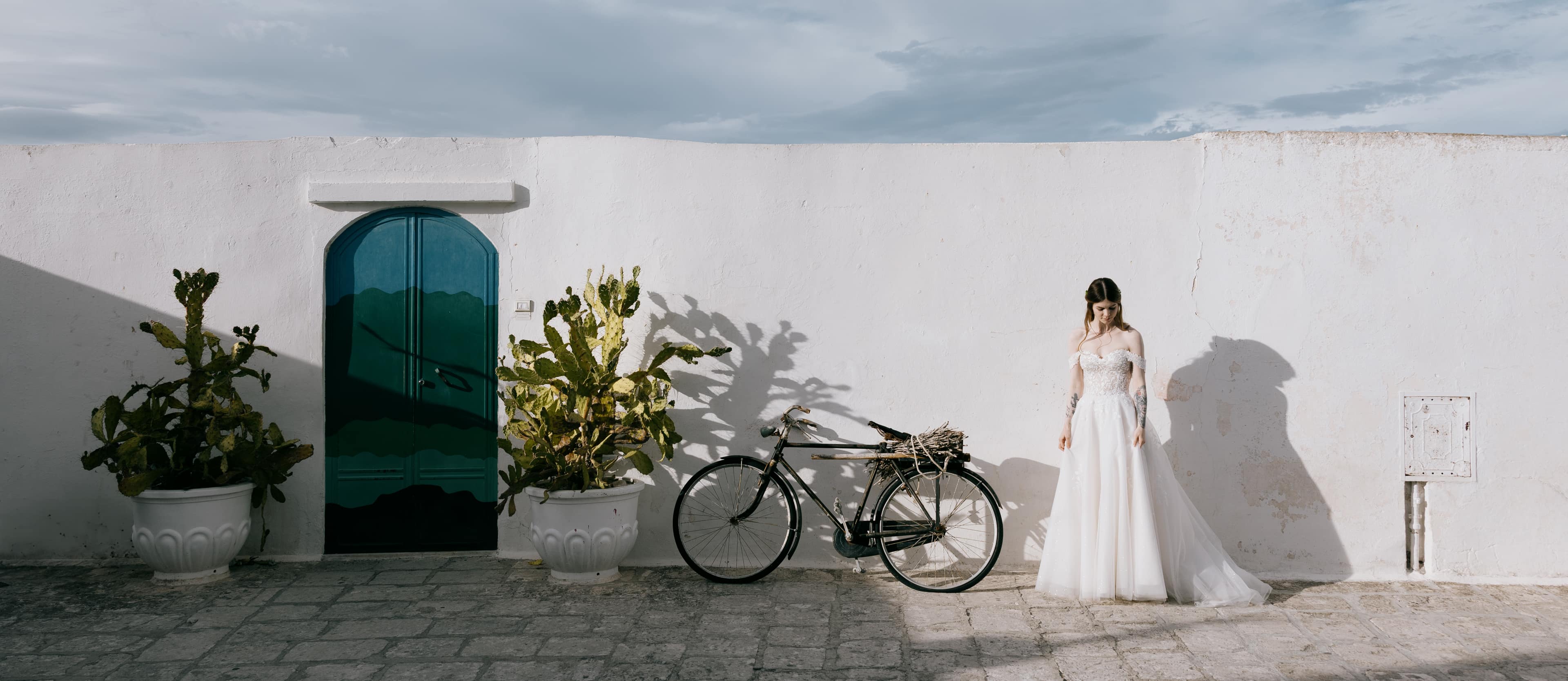 Bride standing in a white-walled Ostuni corner with a bicycle and cactus.