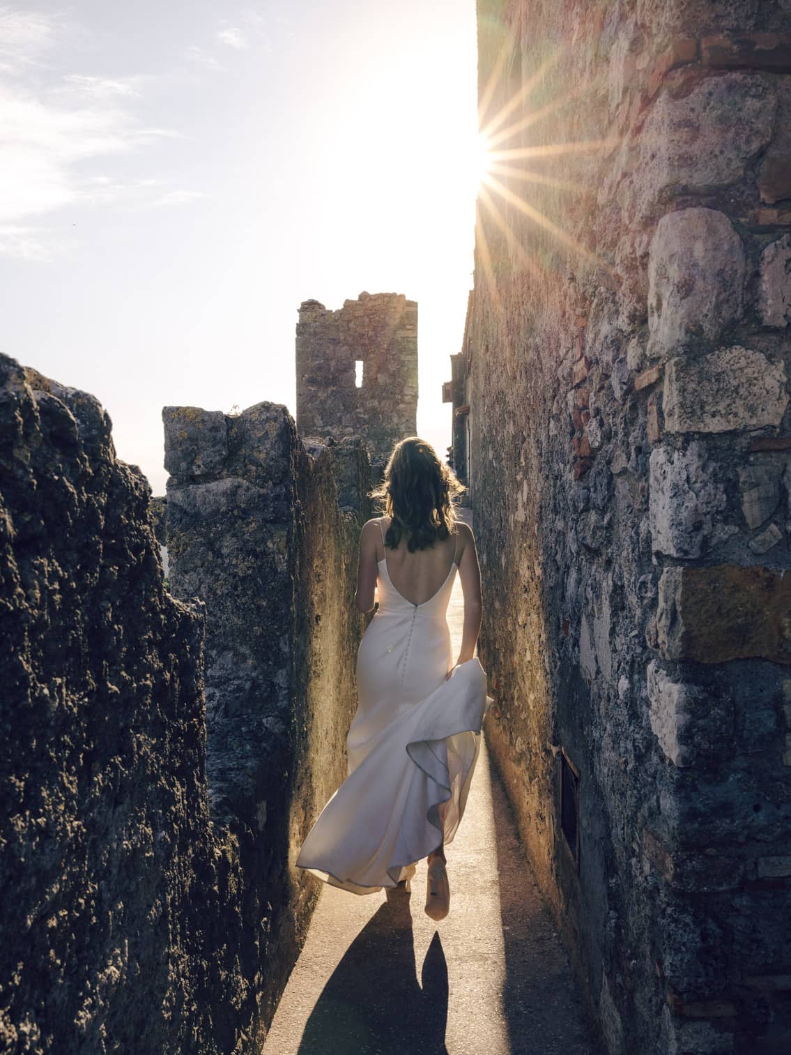Bride walking along ancient castle walls in Tuscany, backlit by a warm sunset.